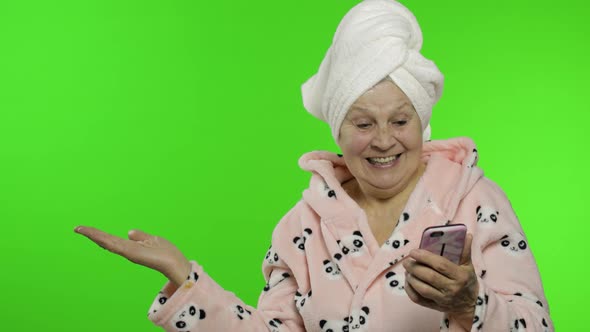 Elderly Grandmother in Bathrobe. Old Woman with Smartphone Pointing at Something alt