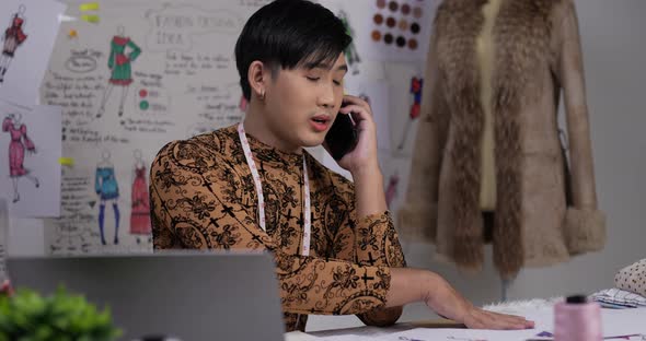 Asian clothing designer working on laptop and talking on mobile in his office alt