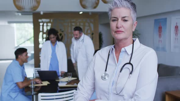 Portrait of caucasian female senior doctor smiling, with colleagues in discussion in background alt