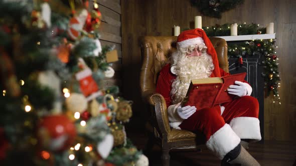 Santa Claus Pulls Out Old Red Silk Album Near Christmas Tree with Toys and Lights alt