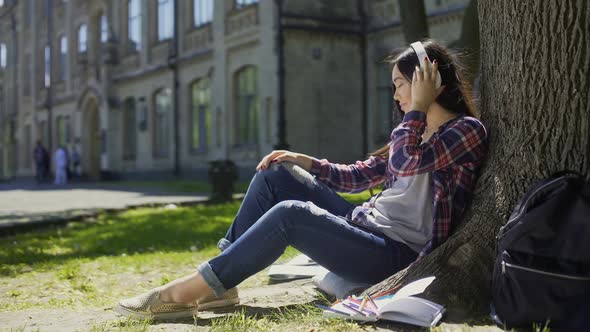 Multinational Young Woman in Headphones Sitting Under Tree, Listening to Music