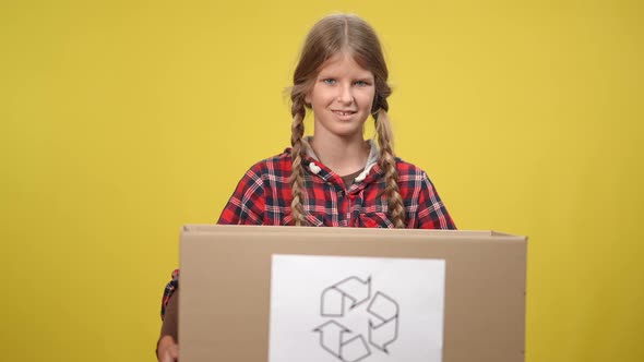 Smiling Teenage Girl Holding Cardboard Box with Recycle Symbol Looking at Camera alt