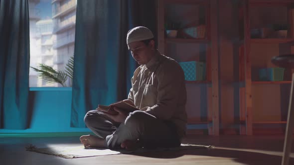 Dark Shot Portrait Muslim Man in Traditional Clothing Reads a Book at Home alt