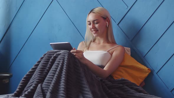 Girl with Headphones is Sitting on Bed Covered with Blanket and Talking Video Link on Electronic alt