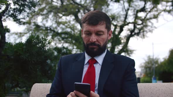 Serious Bearded Man in an Expensive Suit Typing Something on His Phone While Sitting on a Park Bench alt