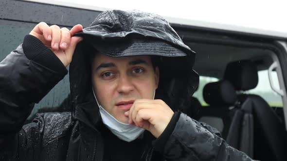 A Man in a Black Jacket in a Heavy Rain Puts a Medical Mask on His Face and Waves His Hand alt