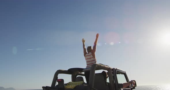 Happy caucasian gay male couple in car raising arms and waving on sunny day at the beach alt