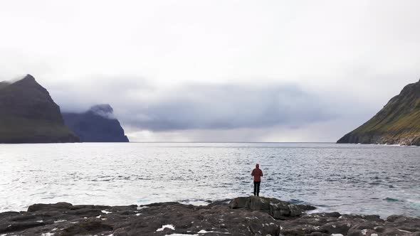 Aerial View of Unrecognizable Woman Stands on Rock Cliff Looking at Wilderness Background of Faroe alt
