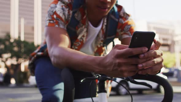 African american man in city, sitting on bike in street using smartphone alt