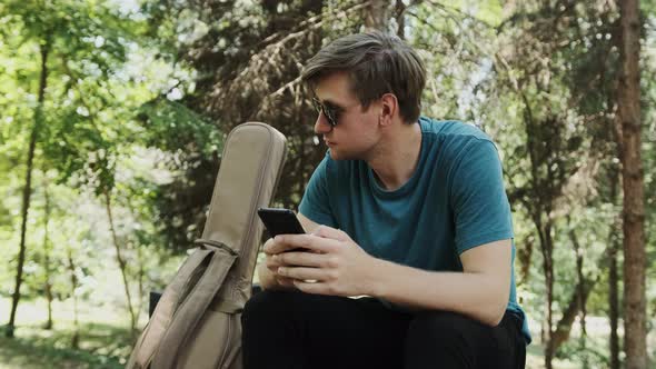 Young Man Sitting on Bench in Park While Is Chatting with His Friends alt