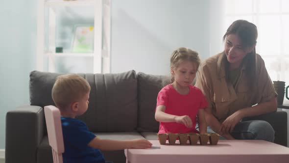Young Woman Watches How Children Plant Seeds of Home Plants alt