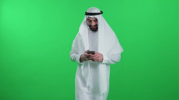 Arab Man in a White Kandura on the Green Background Walks Using a Smartphone and Surf the Internet alt