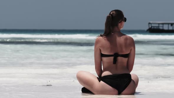 Young Woman in Black Bikini Tans in Lotus Pose on Paradise Sandy Beach By Ocean alt