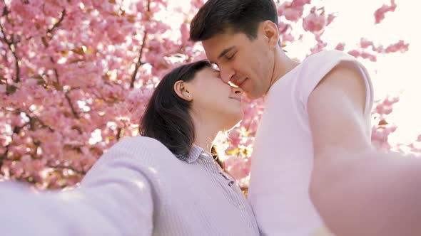 Happy young couple taking selfie by mobile phone near the Blooming tree in summer day. alt