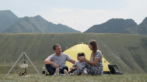 A Happy Family with a Child Is Resting Together in Front of a Tent in the Mountains and Drinking Tea alt