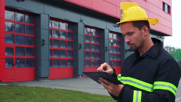 A Young Firefighter Works on a Tablet - a Fire Station in the Background alt