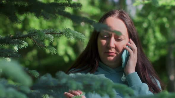 Woman is Calling on Mobile Phone Standing Behind the Green Trees in Park