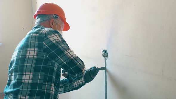 An Elderly Male Worker in a Helmet Mask and Goggles Breaks the Wall with a Chisel and Hammer to Wire