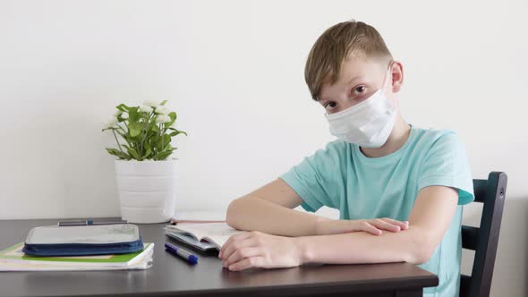 A Young Boy in a Face Mask Looks Up From a School Notebook To the Camera and Points at It at a Table alt