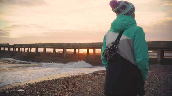 Attractive Young Woman in a Warm Jacket Walking on the Stone Beach at Sunset alt