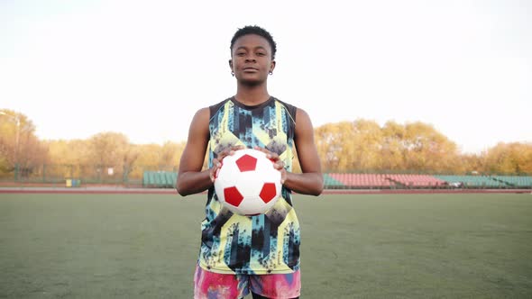 A Serious Young Black Girl in a Sleeveless Tshirt Stands on a Soccer Field and Holds a Ball in Her alt