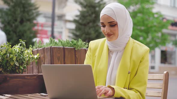 Smiling Young Muslim Woman Wearing Headscarf Sits in Cafe and Uses Laptop alt