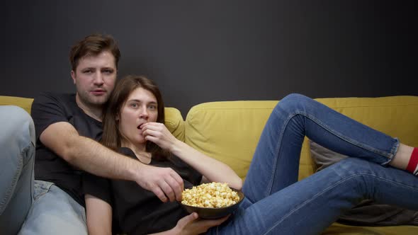 Attractive Young Couple Sitting on Sofa with Popcorn Watching Movie on TV