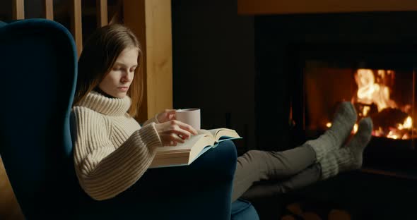 Lady Reads Book and Drinks Tea Relaxing at Fireplace in Armchair at Winter Night