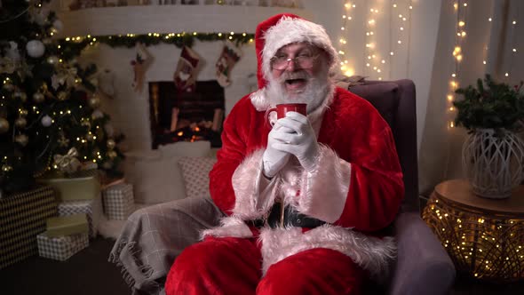Traditional Santa Claus Is Sitting in a Chair Is Drinking a Warm Drink From a Cup Near Christmas