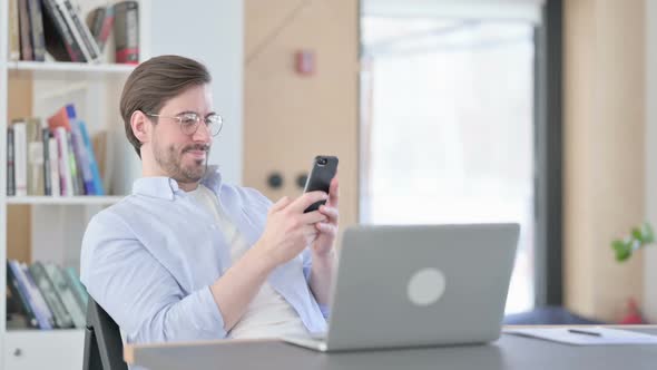 Man in Glasses with Laptop Using Smartphone in Office alt