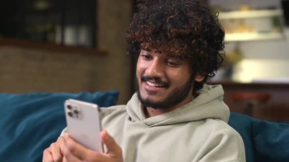 Focused Bearded Indian Man Sitting on Sofa at Home and Holding Smartphone alt