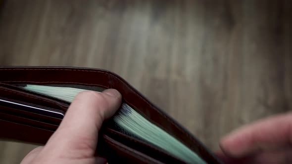 A man's hands open a wallet filled with 100 dollar bills and pass one bill to someone. alt
