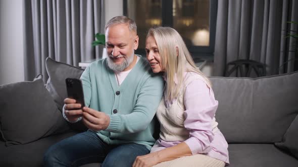 Elderly Couple Sits on a Sofa in the Living Room and View Photos on a Smartphone Family Day Positive alt
