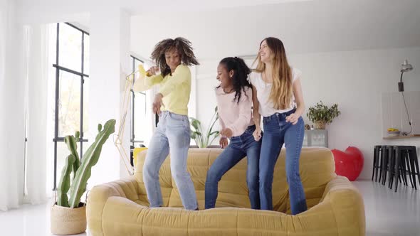 Three Funny Female Friends Dancing Standing on the Sofa Indoors alt