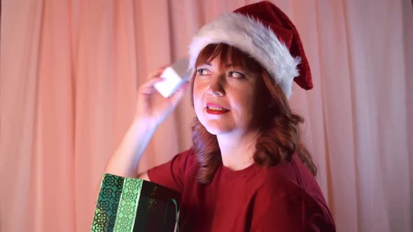 Woman wearing santa hat is smiling and shaking the Christmas present alt
