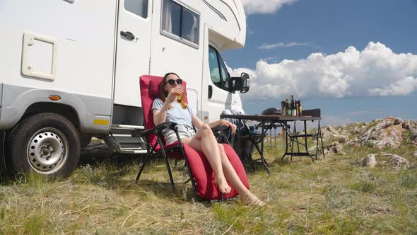 Camper Rv Woman Sitting On Folding Chair Near Motor Home and Drinking Juice alt
