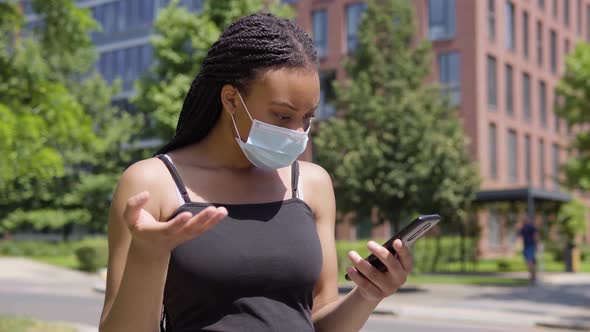 A Young Black Woman in a Face Mask Looks at a Smartphone and Gets Upset - an Office Building alt