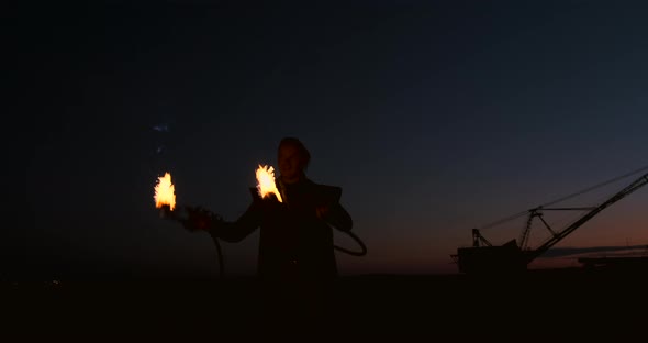 A Man in a Raincoat with Two Flamethrowers Lets Out a Fiery Flame Standing at Sunset on the Sand alt