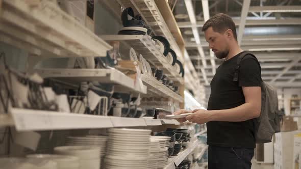 Man is Inspecting Ceramic Plates in Crockery Shop Taking and Putting Back alt