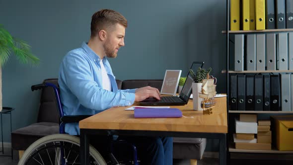 Motivated Disabled Person Freelancer is Working with Laptop Typing Sitting in Wheelchair at Office alt