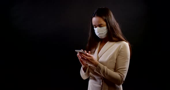 Brunette Woman with Medical Mask on Face During Coronavirus Pandemic Is Using Cell Phone alt