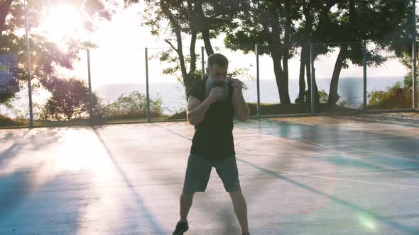Middle Aged Man with Long Gray Beard Boxing with Shadow on Basketball Court During Sunrise alt