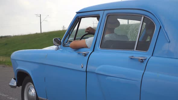 Young Couple in Hats Driving at Blue Retro Car After Rain During Summer Travel. Unrecognizable Pair alt