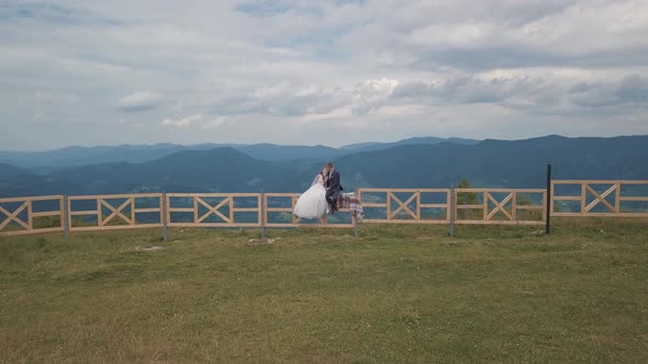 Groom with Bride Sit on the Fence Near Mountain Hills. Aerial Drone Shot alt