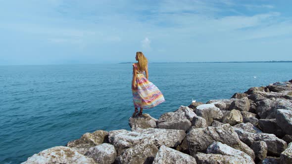 Girl in a Beautiful Dress on Stones Looking at the Ocean alt