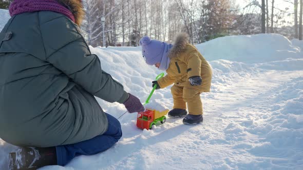 Mom plays with toddler in winter park with toys. Mom loads snow with her palms into a toy truck alt