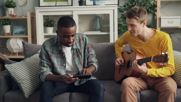 Caucasian Young Man and His African American Friend Are Learning To Play the Guitar Using Tablet alt