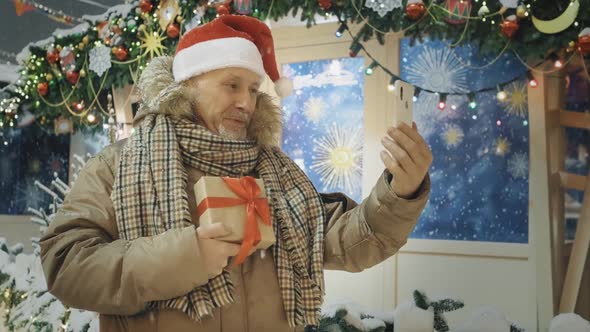 An Elderly Man in a Santa Claus Hat with a Gift Box in His Hands is Talking on the Phone at a