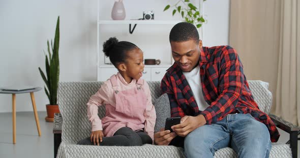 Loving Father Sits on Couch with Little Daughter Preschool Afro American Girl with Smart Phone alt