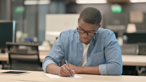Hardworking Young African American Man Writing on Paper in Office alt